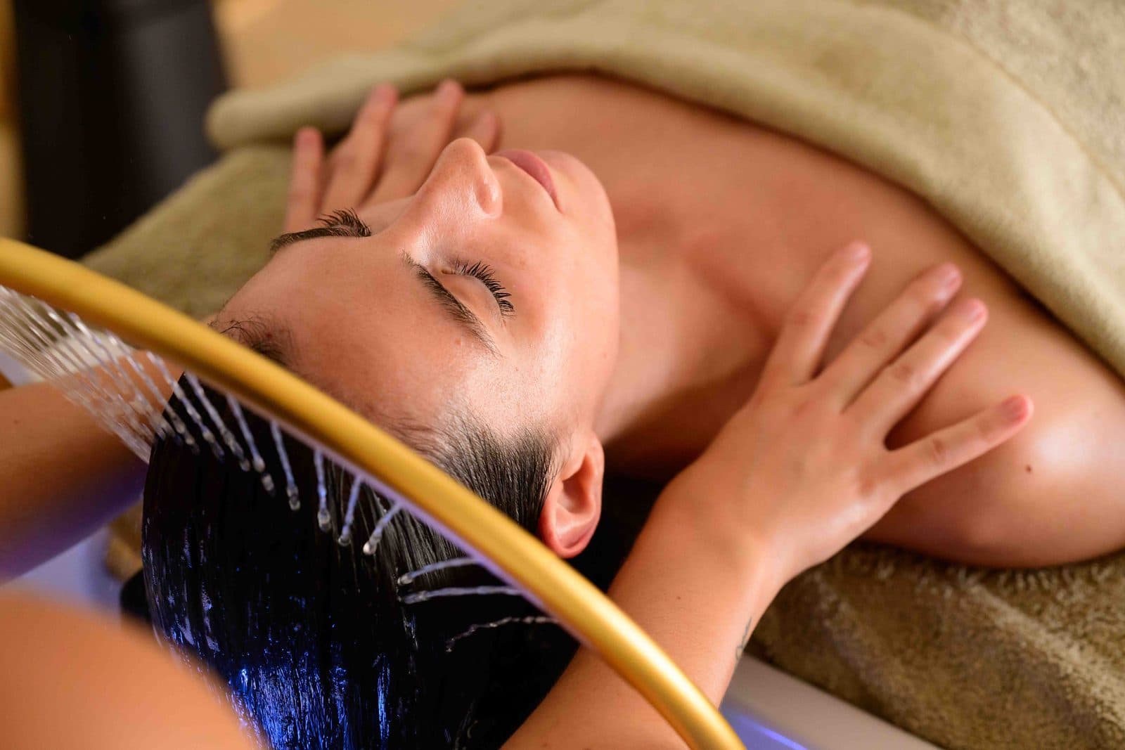 Head spa treatment in a salon, client lying with eyes closed, hair rinsed under a fine jet of water above a basin